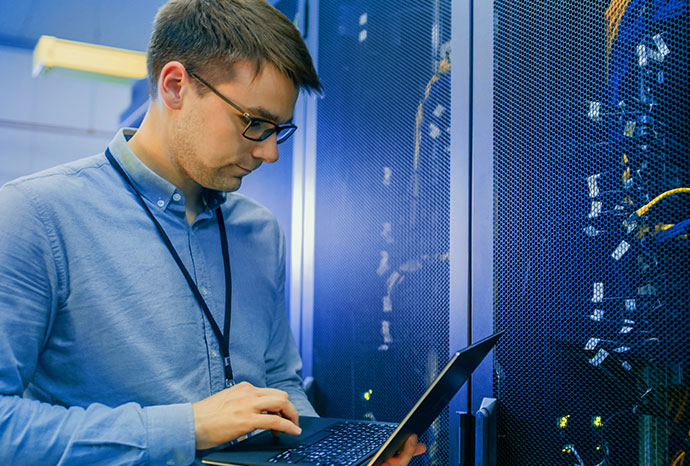 Data Center IT Engineer stands before server rack doing routine maintenance and diagnostics using a laptop